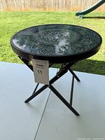 Photo of the assembled round black metal folding plant stand with decorative green mandala patterned glass top on a table outdoors.