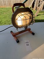 Front view of the shop light showing the protective metal cage around the bulb, the orange metal frame, and the handle on top.