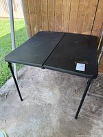 Square black folding table set up outdoors on concrete with metal legs visible and a label on top.