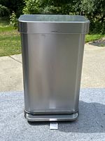 Front view of a stainless steel rectangular kitchen trash can featuring a flat lid and foot pedal, outdoors in daylight.