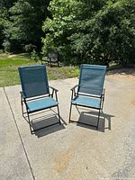 Two teal folding lawn chairs placed outdoors on a paved surface, showing front view and overall condition.