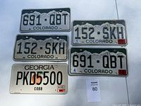 Five vintage license plates arranged on a flat surface: four from Colorado with mountain design and one from Georgia Cobb County.
