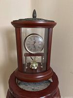 Front view of cylindrical wood and glass mantel clock showing dial with Roman numerals and brass-tone pendulum