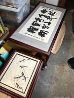 Photo of two Chinese art wall hangings stacked, showing calligraphy framed with a dark wood frame.