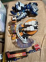 Lot overview showing three Transformers toys on paper background: Optimus Prime truck and trailer, orange-yellow horned robot, blue-gray dinosaur robot.