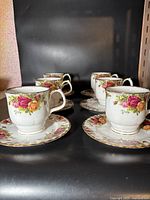 Six Royal Albert Old Country Rose bone china coffee cups with matching saucers arranged on a shelf, showing floral pattern and gold trim.