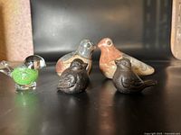 Six bird figurines arranged on black surface: two large painted ceramic/clay, two black cast metal, one green glass, one black with clear wings.