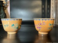 Two Chinese teacups side by side showing mustard yellow glaze with floral and bamboo painted decorations