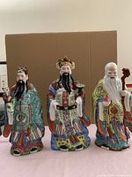 Three Chinese porcelain figurines in traditional robes, standing side by side against a cardboard background