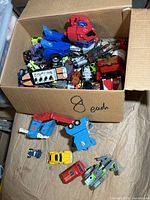 Overview of cardboard box filled with loose Transformers vehicle and robot parts, including red and blue Optimus Prime cab and assorted accessories