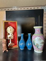 Overview photo showing pink porcelain vase, blue vases, ivory carving in display box, and wooden figurine on black surface
