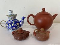Four small Chinese teapots including one blue enamel, one brown clay, two stone in reddish and light brown colors arranged on a white surface.