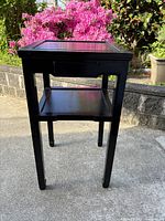 Full front view of the black wooden side table with drawer and lower shelf showing the tapered legs and open drawer.