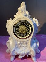 Front view of the white ceramic mechanical clock with ornate curving details and round black and gold dial visible.