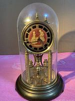 Front view of German mechanical anniversary clock under glass dome highlighting dancing couple clock face, Roman numerals, brass features, and rotating pendulum balls.