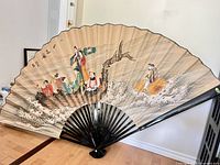 Full view of large hand painted Chinese wall fan showing entire fan with colorful figures and waves, set against wall with wood floor.