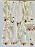 Full view of jewel lot laid out on white background showing seven faux pearl multi-strand necklaces with a variety of colored and clear rhinestone embellishments plus one pair of clip-on earrings.