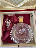 Empty crystal Louis XIII decanter with gold-tone collar and fleur-de-lis motifs alongside its stopper on burgundy fabric lining