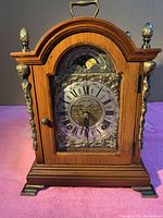 Front view showing wooden arched case with brass ornaments and Roman numeral dial with moon phase above.