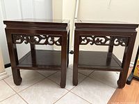 Front view of two Chinese wood carving side tables showing apron carving and lower shelf on tiled floor.