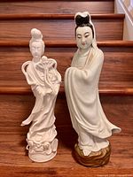 Both porcelain Guanyin statues shown from front on wooden stairs, one taller and one smaller.
