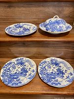 Four pieces of blue and white porcelain with bird and floral patterns displayed on a wooden surface: three plates and one covered dish with lid.