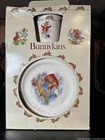 Front view of the ceramic plate showing a colorful Bunnykins illustration of rabbits on a boat, with surrounding decorative rabbits on the packaging.