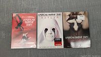 Front view of three American Horror Story DVD box sets Seasons 1-3, showing the front cover artwork: Season 1 with red background and falling woman, Season 2 with white background and crying nun, Season 3 with dark cover and two women.