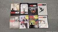 Eight DVD box sets for Mad Men TV series Seasons 1-7 displayed on a gray surface, showing front covers with various distinctive artwork.