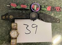 Three vintage wristwatches arranged on a surface with a sheet marked '39'. Two watches show fabric and leather straps; the third has a metal expandable band.