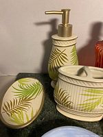 Photo of ceramic bathroom set items including soap dish, lotion dispenser, and divided container featuring green and brown leaf patterns on cream ribbed base.