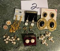 Photo of a set of eight assorted earrings displayed on a table, showing various styles including gold-tone dangle, pearl clusters, and black stone designs. Some earrings are on original branded cards.
