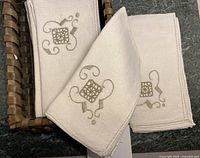 Set of 12 beige-colored dinner napkins with decorative embroidery in a brown basket on dark tabletop.