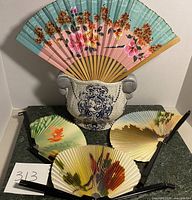 Four decorative folding fans displayed in and around a vintage blue crackle ceramic vase with ornate handles and blue crest design on front.