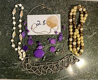 Four necklaces displayed on a green marble surface: white bead necklace with floral oval beads, purple bead necklace with silver chain, silver-tone rhinestone necklace, and multi-strand faux pearl necklace.