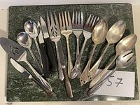 Overview of all serving utensils displayed on dark green marble slab, various spoons, forks, knives including black-handled knives and cheese slicer