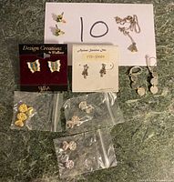 Photo showing multiple pairs of small earrings including colorful butterflies, floral shapes, and a small chain necklace with bell pendant arranged on a surface.