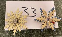 Pair of two snowflake-shaped brooch pins placed on numbered white card showing front detail of enamel and rhinestone design.
