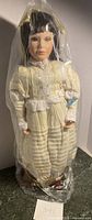 Full view of the tall porcelain doll Jennifer dressed in ivory dress with lace trim, dark brown wig, sealed in plastic wrapping.
