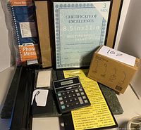 Photo of assorted office supplies including two Adams Phone Message pads, 3-pack of multipurpose certificate frames, black AAD calculator, yellow label sheet, boxed desk lamp and two small white plastic items on green background