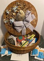 Woven sewing basket showing decorative lid with dried flowers, fabric bird, and gray ribbon bow. Opened basket revealing various sewing notions inside including pincushion, scissors, needles, and knitting needles.