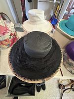 Black ladies hat with lace and veil on a table, white ladies hat in background, floral hat box also visible.