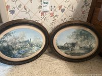 Photo showing two oval framed prints leaning against a wall with floral wallpaper, titles not visible but frames and prints clearly shown.