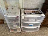 Two plastic drawer units side by side, one smaller with four drawers and one larger with three drawers, all semi-transparent white plastic. Units contain various household items visible inside drawers.