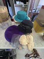 Four ladies hats including teal wide-brimmed, black rounded with bow, purple velvet, and cream fur with netting, displayed on a surface with two hat boxes nearby.
