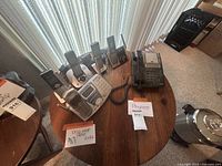 Lot of six phones on round wood table near window: five cordless phones on bases and one large Panasonic office phone with keypad and display.