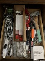 Drawer view showing assorted kitchen utensils including metal whisks, cheese graters, wooden spoons, and various single wrapped utensils.