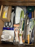 Drawer containing assorted kitchen utensils including scissors, tongs, egg slicer, food slicer, and whisks, many in original packaging indicating some new items.