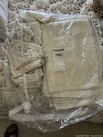 Image showing several cream and off-white vintage table linens folded and wrapped in plastic, including a large tablecloth with visible embroidered patterns and scalloped edges.