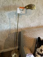 Full view of the brass floor lamp standing beside a couch, showing the shell-shaped lamp shade pointing forward and the round base on the floor.